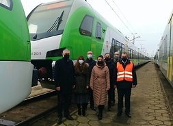 Grupa siedmiu osówb (dwie kobiety i pięciu mężczyzn) stoi między składami nowych pociągów. Wszyscy mają maseczki