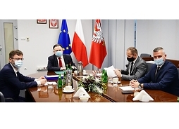 Czterech panów w maseczkach ochronnych, siedzą wokół stołu, przed nimi dokumenty, z tyłu w stojaku flagi Polski, UE i Mazowsza.