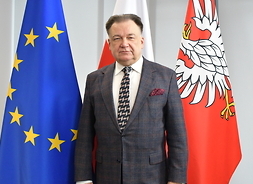 Marszałek Struzik w garniturze stoi na tle flag UE, Polski i Mazowsza. Pozuje do zdjęcia