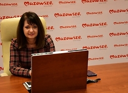 Członek zarządu Janina Ewa Orzełowska siedzi przy stole konferencyjnym. Przed nią widać otwarty laptop. W tle jest ścianka z logiem mazowsza