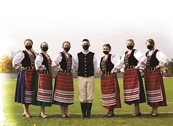 Grupa tancerzy w strojach ludowych z maseczkami ochronnymi na twarzy pozuje do zdjęcia w plenerze.