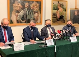 Czterech panów w garniturach i maseczkach siedzi za stołem prezydialnym, za nimi na ścianie trzy obrazy pędzla Malczewskiego z kolekcji muzealnej.