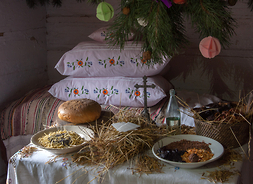 izba wiejska, na stole, pokrytym białym obrusem leżą w naczyniach potrawy, w rogu stoi duża zielona choinka przyozdobiona ozdobami z papieru