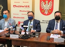 Trzech panów w garniturach i maseczkach ochronnych siedzi w rzędzie za stołem, za nimi baner z logotypami samorządu Mazowsza.