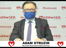 Męzzczyzzna w garniturze, okularach i maseczce ochronnej z mikrofonem siedzi za stołem, za nim banner z logotypami samorządu Mazowsza..
