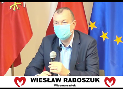 Męzzczyzzna w garniturze i maseczce ochronnej z mikrofonem siedzi za stołem, za nim flagi Polski, UE i Mazowsza.