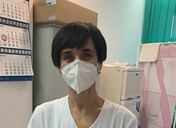 Kobieta w lekarskim fartuchu i maseczką na twarzy trzyma przed sobą dokument potwierdzający szczepienie na Covid-19.