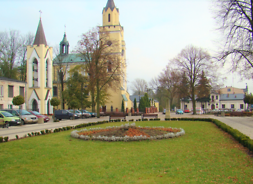 Plac z trawnikiem i deptakiem, z tyłu za nim zespół budynków kościelnych.