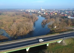W dużym planie widok miejscowości, w oddali wiele budynków i płynąca rzeka - na pierwszym planie wiadukt na którym jedzie samochód osobowy.