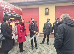 Obok wozu strażackiego stoi członek zarządu Janina Ewa Orzełowska. Wokół niej w pewnej odległości zgromadzeni są inni uczestnicy wydarzenia. Wszyscy na nią patrzą