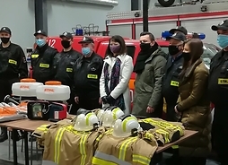 Członek zarządu Janina Ewa Orzełowska stoi wśród strażaków. Przed nimi na stole rozłozone są elementy stroju strażackiego oraz podarowny sprzęt