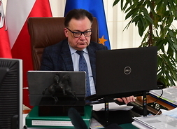 Marszałek Adam Struzik siedzi za biurkiem w swoim gabinecie. Wpatruje się w monitor, obok leży otwarty ekran. W tle widać flagi Unii Europejskiej, Polski i flaga z godłem województwa