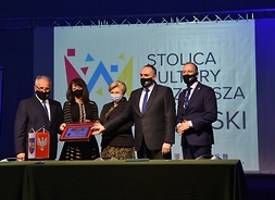 Elżbieta Lanc i Janina Ewa Orzełowska wraz z trzema przedstawicielami powiatu stoją za stołem i wspołnie trzymają klucz do stolicy kultury Mazowsza