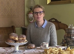 Kobieta w okularach siedzi przy stole, na którym stoi naczynie z pączkami, talerz z faworkami oraz filiżanka.