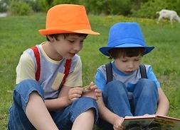 Dwóch chłopców w letnich kapeluszach siedzących na łące i czytających książkę.