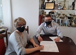Mężczyzna i kobieta w maskach siedzą przy stole, przed nimi leżą dokumenty z planem nowego budynku