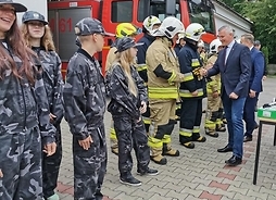 w ciągu stoją strażacy w ubraniach bojowych oraz młodzi ludzie ze strażackich drużyn młodzieżowych, przyjmują gratulacje od wicemarszałka Rafała Rajkowskieggo