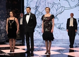 Trzech mężczyzn i dwie kobiety w strojach wieczorowych występują na scenie. W tle dekoracje z motywami roślinnymi
