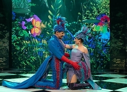Dwoje artystów w kostiumach scenicznych klęczy na przeciwko siebie, patrząc sobie w oczy i trzymając się w objęciach. W tle dekoracja sceniczna przedstawiająca rośliny i motyle.