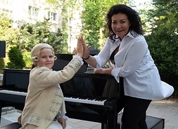 Dyrektor WOK Alicja Węgorzewska przybija dłoń do dłoni chłopca w peruce i stroju z epoki mozartowskiej. Chłopiec siedzi przy fortepianie.