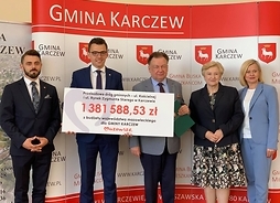 Trzech mężczyzn i dwie kobiety stoją na tle ścianki promocyjnej gminy Karczew. Jeden z mężczyzn trzyma symboliczny czek z wpisaną kwotą dofinansowania.