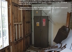 Wnętrze jednego z pomieszczeń przed remontem. Widać zniszczone drewniane ściany oraz rozsypane na podłodze kawałki zdartej płyty. W rogu stoi stara, metalowa szafka.