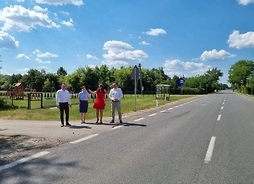Cztery osoby stoją przy drodze z zaniedbanym, nieutwardzonym poboczem