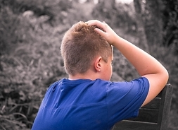 Chłopiec siedzący na ławce, bokiem zwrócony do obiektywu, trzyma się za głowę w geście rezygnacji