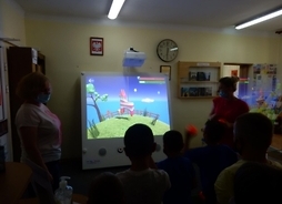 Dzieci w bibliotece wpatrują się w wyświetlany ekran, na którym widać kreskówkę - konia na łące