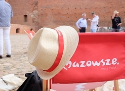 Na pierwszym planie tył leżaka z napisem Mazowsze serce Polski, na brzegu leżaka powieszony słomkowy kapelusz z tasiemką logo Mazowsza