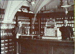 czarnobiała stara fotografia przedstawiająca wnętrze dawnej apteki. Przy ladzie stoją dwaj aptekarze