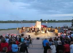 Scena zbudowana na piasku nad Wisłą, na niej kilkunastu śpiewających aktorów, płonące pochodnie