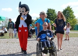 Mama popycha wózek inwalidzki z niepełnosprawnym nastolatkiem, obok aktor przebrany za myszkę Miki