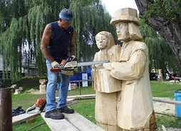 Artysta piłą mechaniczną obrabia rzeźbę dwóch postaci naturalnej wielkości w wielkim kawałku drewna