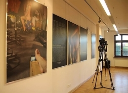Widok na wystawę we wnętrzach muzeum – duże fotosy na ścianie. Statyw fotograficzny na środku