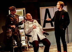Scena ze sztuki przedstawiająca przestraszonego mężczyznę przesłuchiwanego przez dwóch innych ubranych w mundury