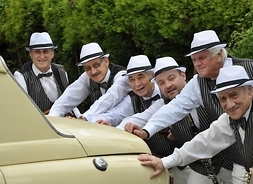 Muzycy ubrani w koszule, kamizelki i kapelusze wzorowane na latach 30. XX w. pchający zabytkowy samochód