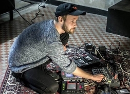 Producent muzyczny siedzący na dywanie nad rozłożonymi wokół elektronicznymi urządzeniami przetwarzania dźwięków i kasetami