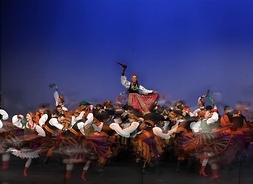 Liczna grupa tancerek i tancerzy w tradycyjnych strojach ludowych, obracających się wokół centralnej grupy, trzymającej podniesioną jedną z pań