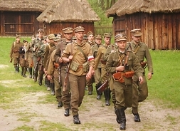 Żołnierze maszerują drogą między drewnianymi wiejskimi chatami pokrytymi strzechą. Mają na sobie mundury i niosą broń