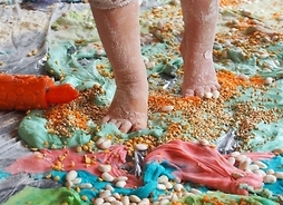 Ubrudzone nóżki dziecka stąpające po rozsypanych fasolkach, ziarnach i wysypanych na kolorowych szmatkach i masach
