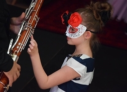 Dziewczynka w sukience w paski i z maską karnawałową na oczach dotyka klarnetu