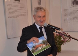 Mężczyzna w garniturze podczas wystąpienia prezentuje książkę. Na okładce dworek - Muzeum Ziemiaństwa w Dąbrowie