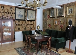 Salon ziemiański ze stołem, zdobnymi krzesłami pluszową kanapą i fotelem oraz kredensem. Na ścianach arrasy i portrety ziemian
