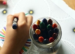 Na zdjęciu widać dłoń dziecka, które kredką maluje rysowankę oraz pojemnik z kredkami