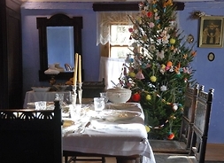 Zdjęcie przedstawia wnętrze wiejskiej chaty udekorowane świątecznie. Na pierwszym planie stoi stół nakryty do świątecznego posiłku