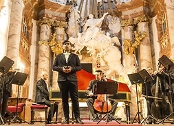 Grupa artystów gra na instrumentach muzycznych, podczas występu. Koncert w barokowych wnętrzach.