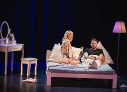 Zdjęcie z przedstawienia, kobieta i mężczyzn rozmawiają ze sobą siedząc na łóżku. Obok łóżka stoi toaletka z lustrem i taboret