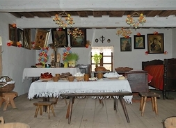 Wnętrze chaty chłopskliej. Na pierwszym planie stoi stół przykryty obrusem. Na stole ustawiono potrawy