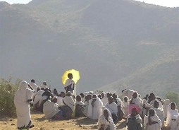 Grupa Etiopczyków w strojach tradycyjnych siedzi na ziemi. Jedna osoba stoi z rozłożonym parasolem slonecznym, druga idzie w kierunku grupy. W tle masyw górski.
